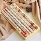 Decorative keyboard with pastel pink, beige, and plaid keycaps, surrounded by soft fabrics, leaves, and pinecones for a cozy aesthetic
