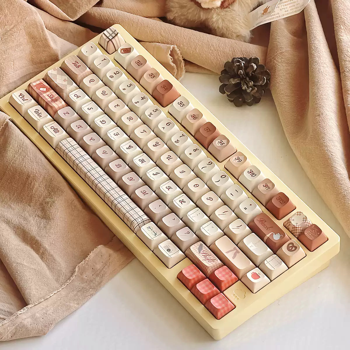 Decorative keyboard with pastel pink, beige, and plaid keycaps, surrounded by soft fabrics, leaves, and pinecones for a cozy aesthetic