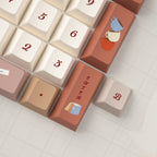 Close-up of pastel pink and beige keycaps with red script lettering and plaid design on a yellow keyboard, surrounded by greenery and soft fabrics